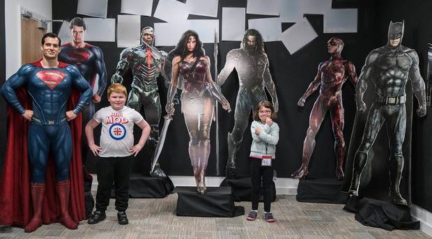 Henry Cavill posando con unos niños de la Fundación Make a Wish y con arte promocional de la Liga de la Justicia